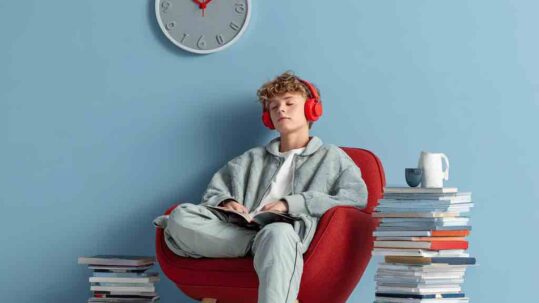 un adolescent assis sur une chaise qui porte un casque rouge sur les oreilles et une seule pile de livres posés à coté de sa chaise. mur bleu pastel et une horloge design au mur --v 7 Job ID: 5abf163d-abed-4b60-9e94-eb00e9b67dea